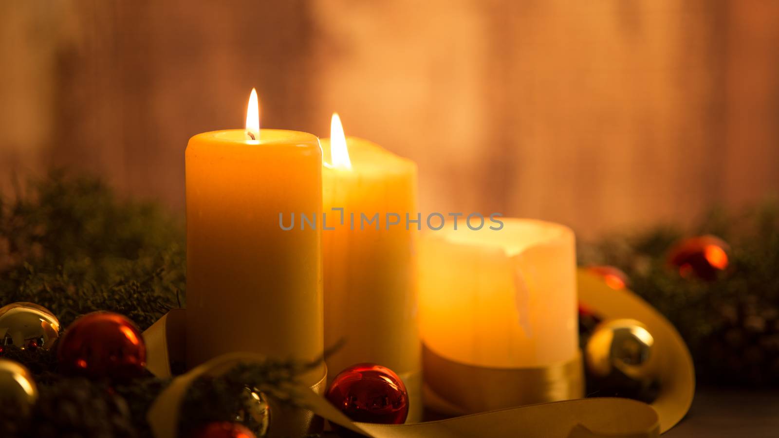 The warmth of the Christmas concept: close up of three candles lit on a dark wooden table and with pine branches, natural pine cones and gold and red bright baubles with a gold satin ribbon