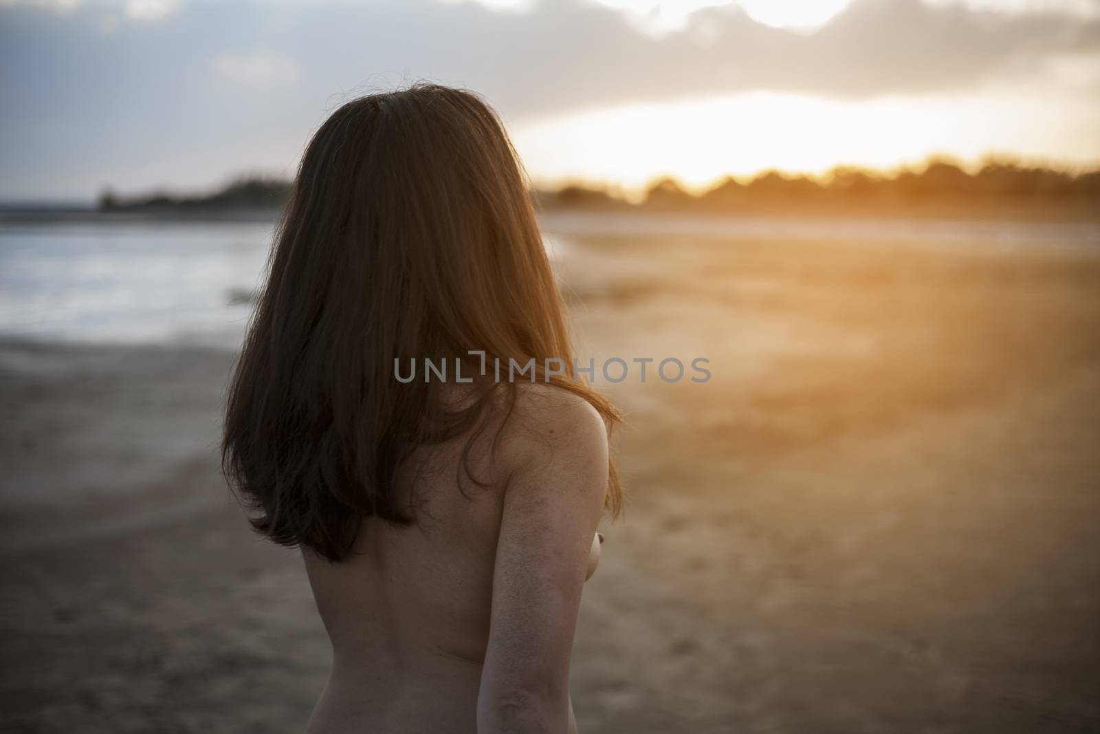 Naked carefree girl on beach with sunset in background. Back view