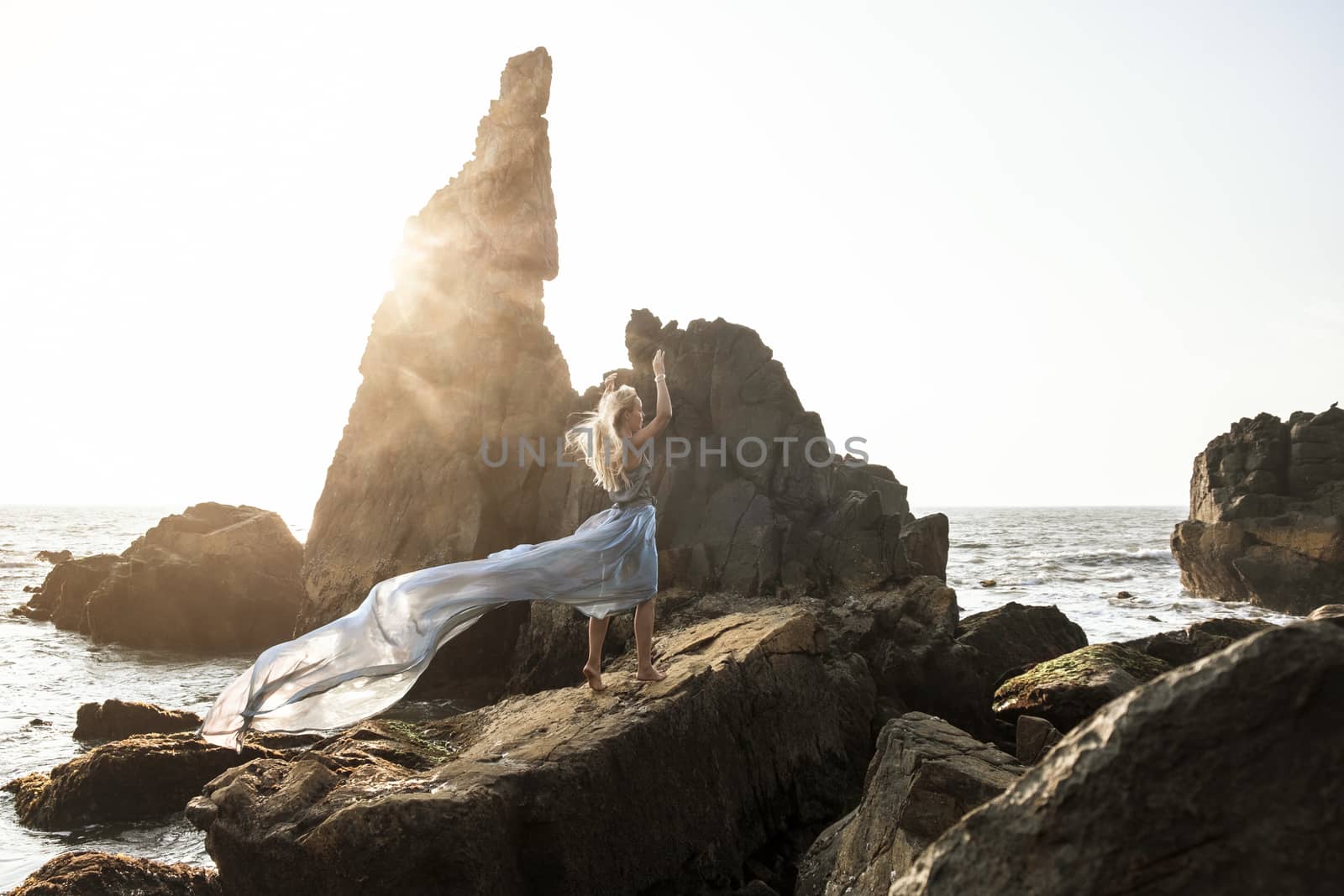 Beautiful European girl in a long dress stands on the rocks by the sea