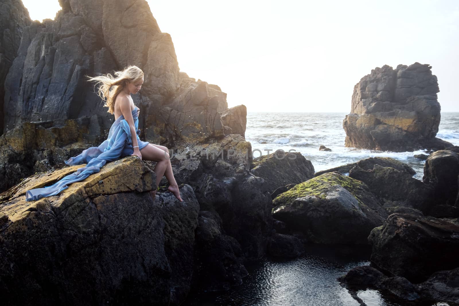 Beautiful European girl in a long dress sitting on the edge of a cliff and looking at the sea