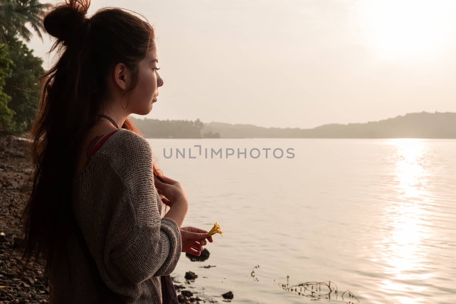 Asian girl stands on the seashore with a flower in her hands, meets the sunset