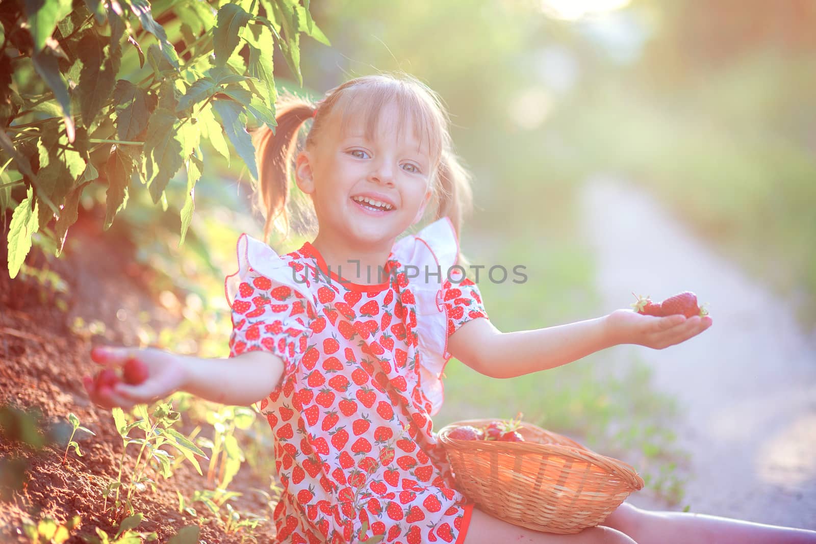 Happy smiling girl in a summer dress sits on the ground in the village and holds strawberry berries in her hands
