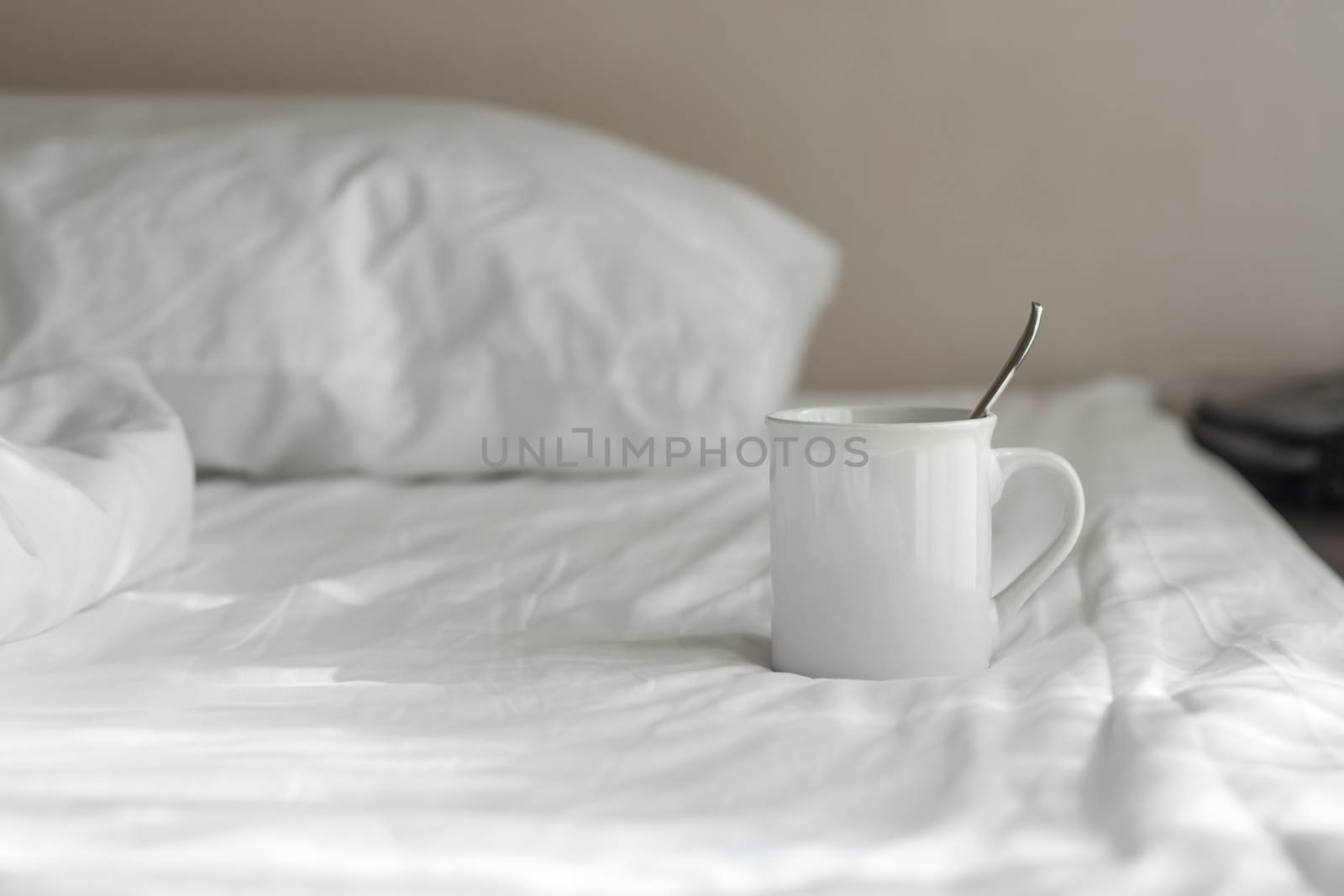 White coffee mug with spoon lay on the bed in the morning.