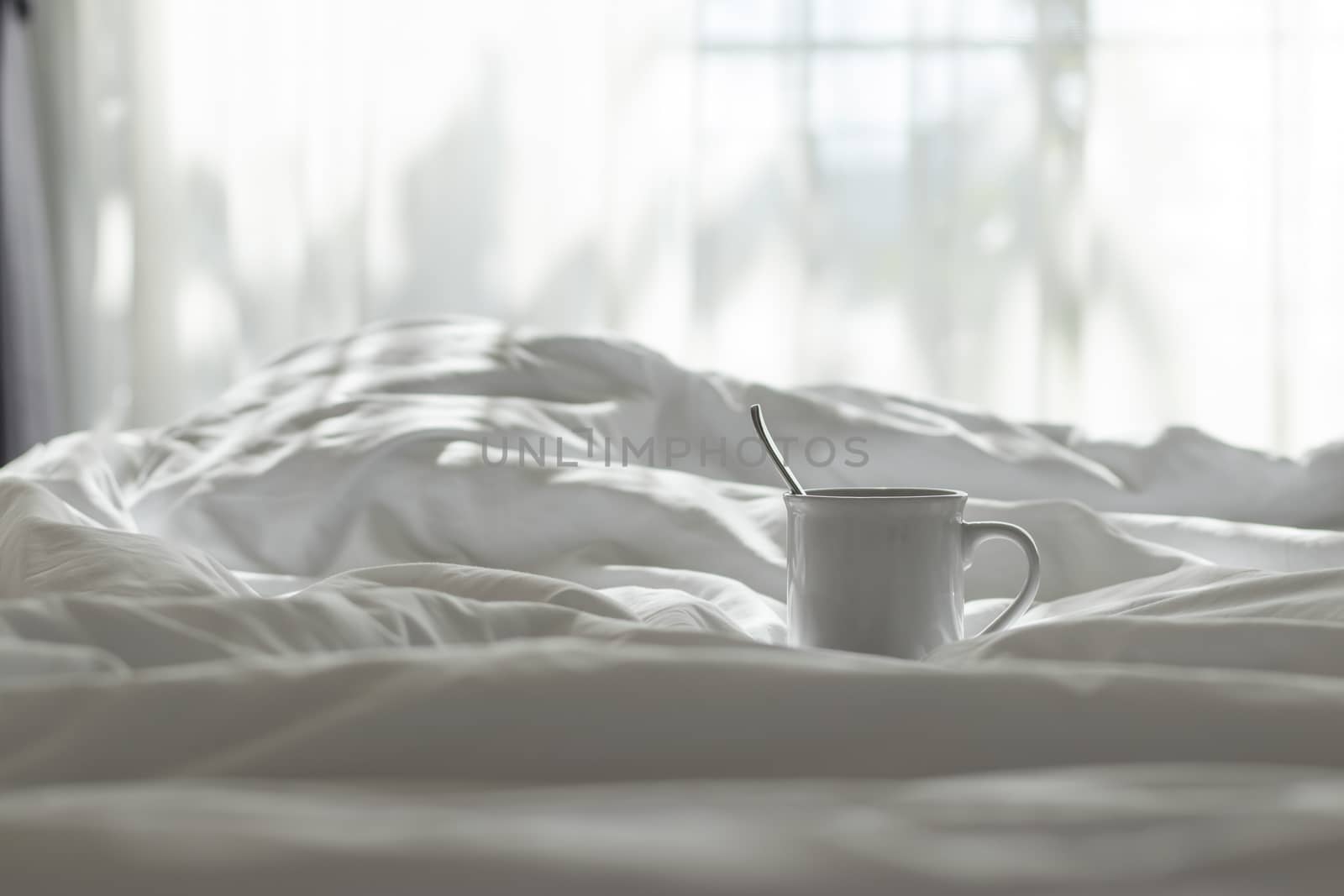 White coffee mug and spoon lay on the bed with the light from the window in the morning. by Eungsuwat