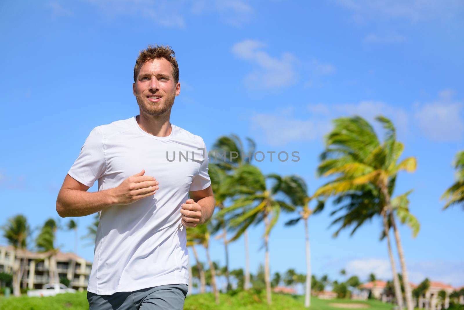 Healthy active man runner running in tropical park. Portrait of handsome young male jogger training cardio going for a run in city park or resort with palm trees in the background in summer.