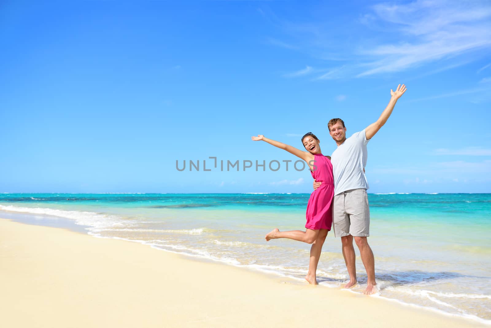Freedom on beach vacation - happy carefree winning couple with arms up showing happiness and fun on paradise beach with perfect pristine turquoise water in sunny tropical getaway