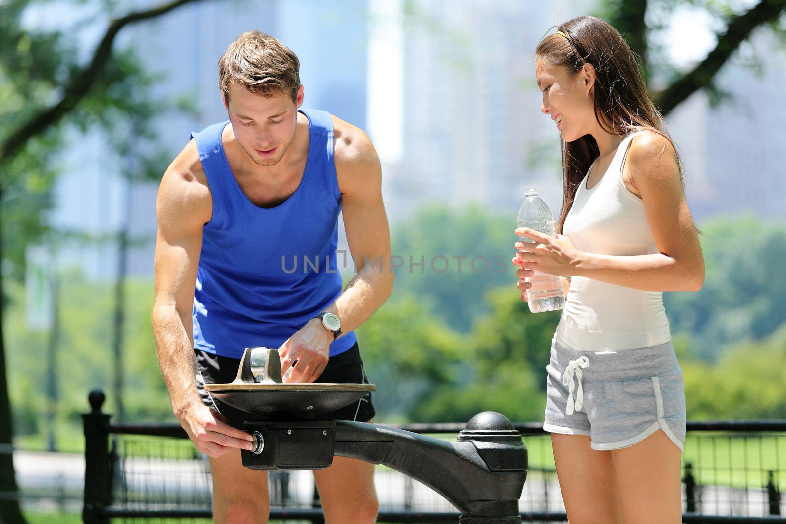 Public park water fountain couple runners drinking. Two young active male and female adults thirsty after run using a drinking fountain in Central Park, New York City, Manhattan in summer, USA.