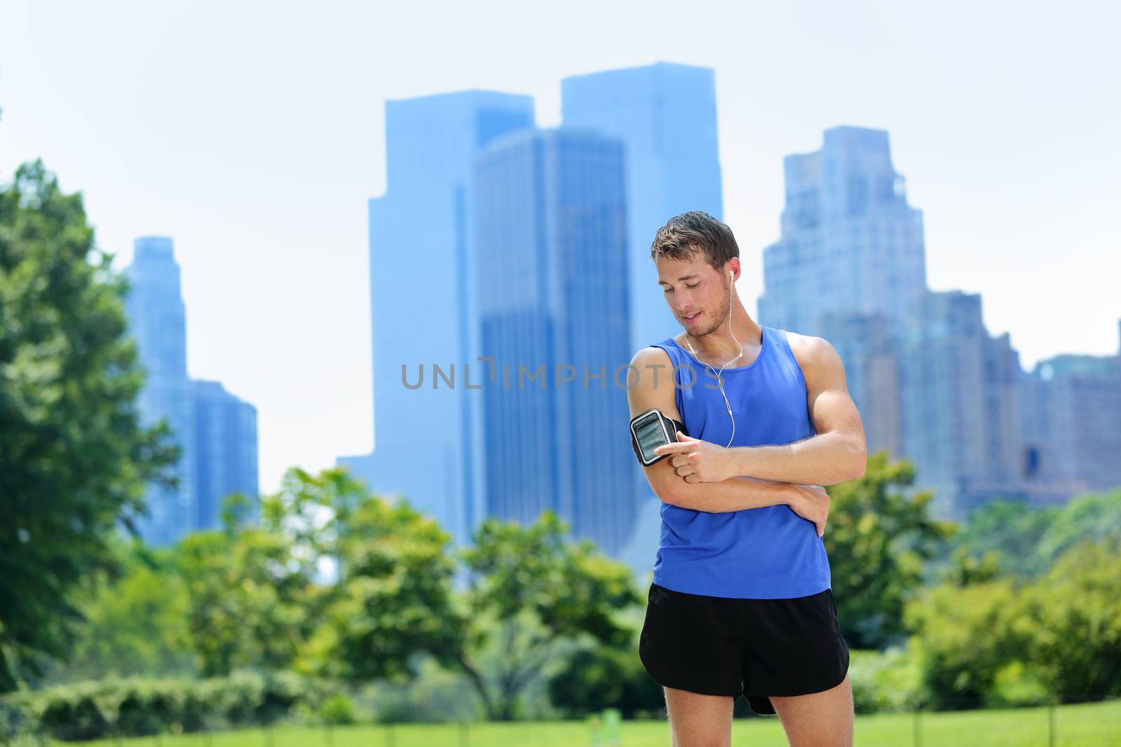 New York City man runner listening music on smartphone. Male adult jogger running using touchscreen on armband for workout in Central Park with urban background of Manhattan's skyscrapers skyline.