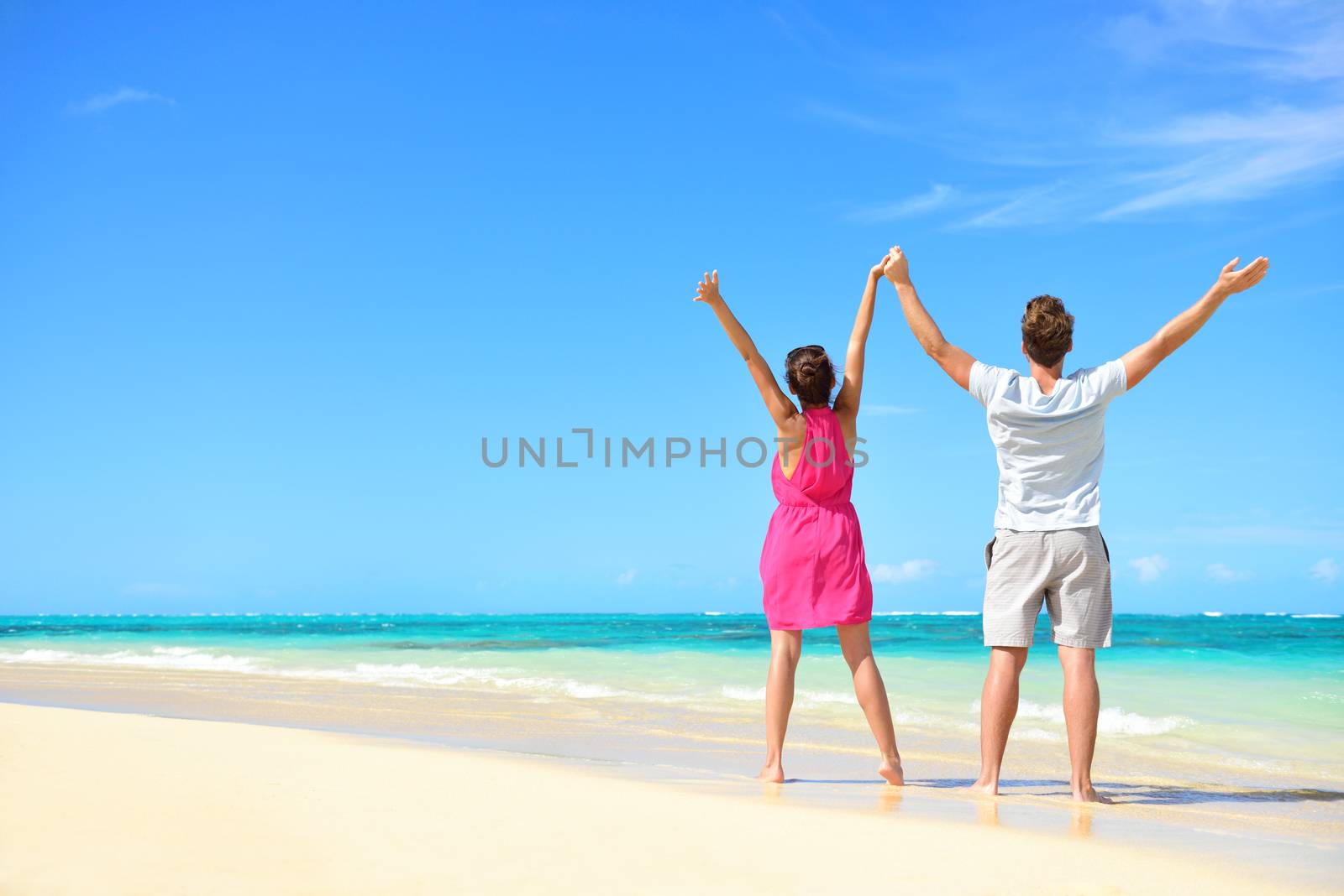 Happy free couple cheering on perfect beach travel holiday. Rear view of unrecognizable young people with arms raised to the sky praising the sun or showing success and freedom during summer vacation.