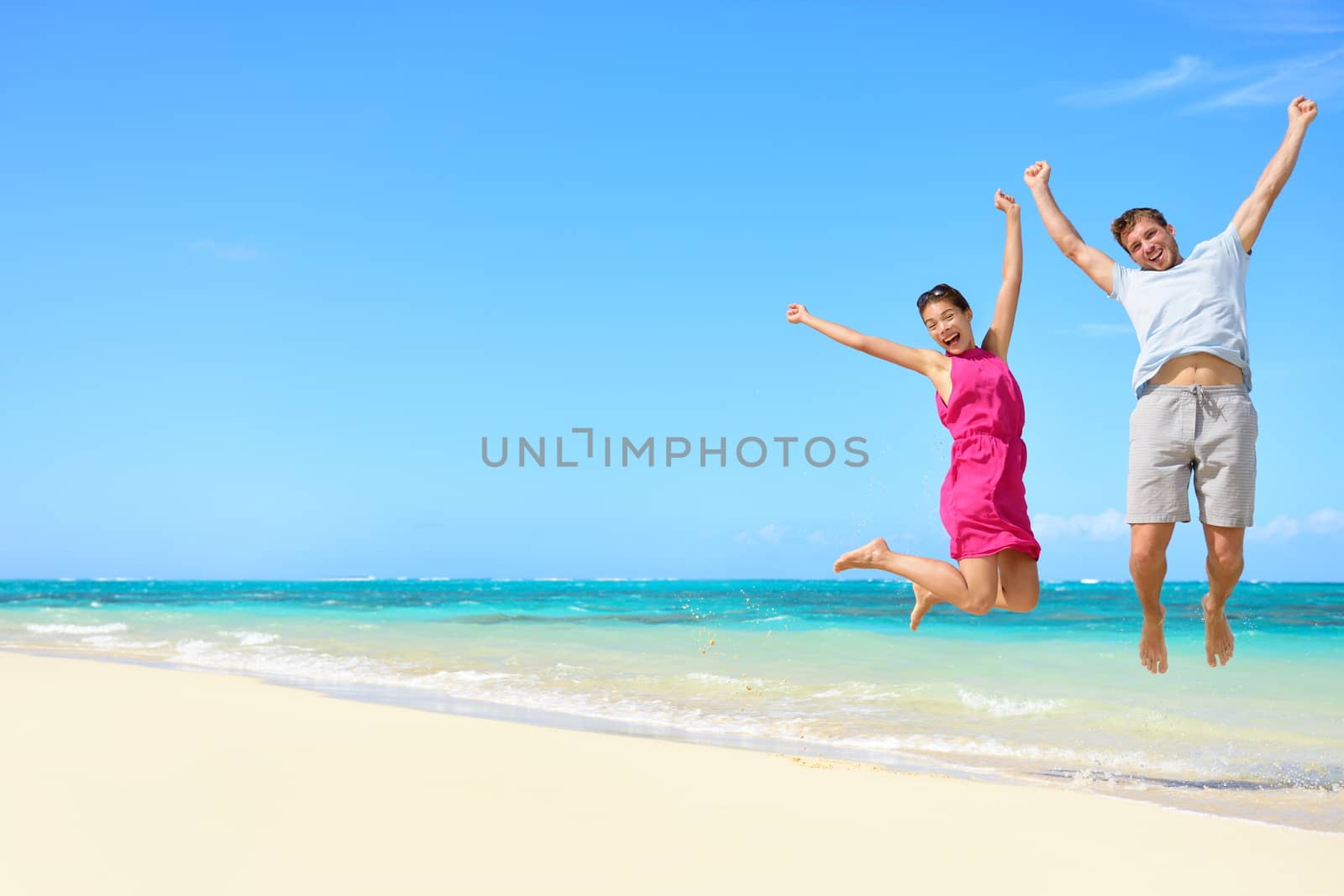 Happy couple tourists jumping on beach vacations. Travel concept of young couple cheering for summer holidays showing success, happiness, and joy on perfect white sand tropical beach under the sun.