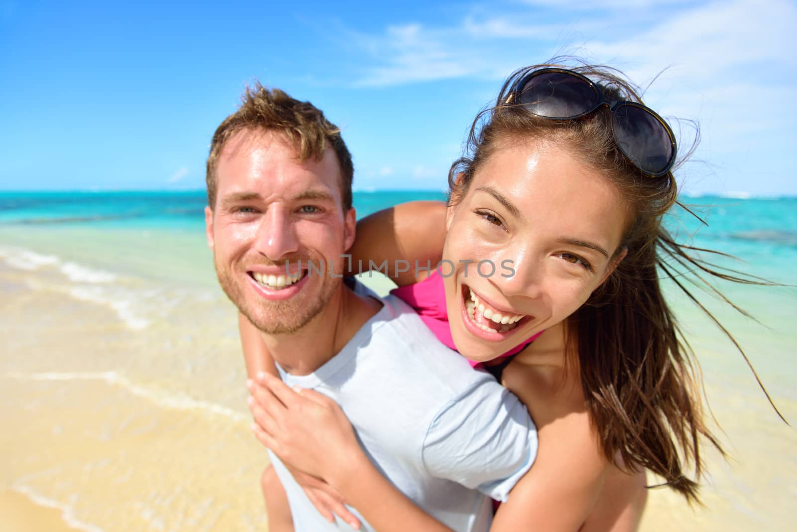 Young couple having fun laughing on beach holidays. Beautiful Asian mixed race woman piggybacking on Caucasian male excited at camera portrait. Multicultural, multiethnic, multiracial people.