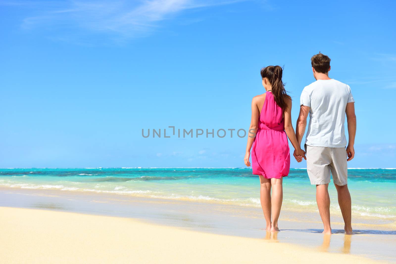 Summer holidays - couple on tropical beach vacation standing in white sand relaxing looking at ocean view. Romantic young adults holding hands in beachwear with pink dress and surf shorts in love.