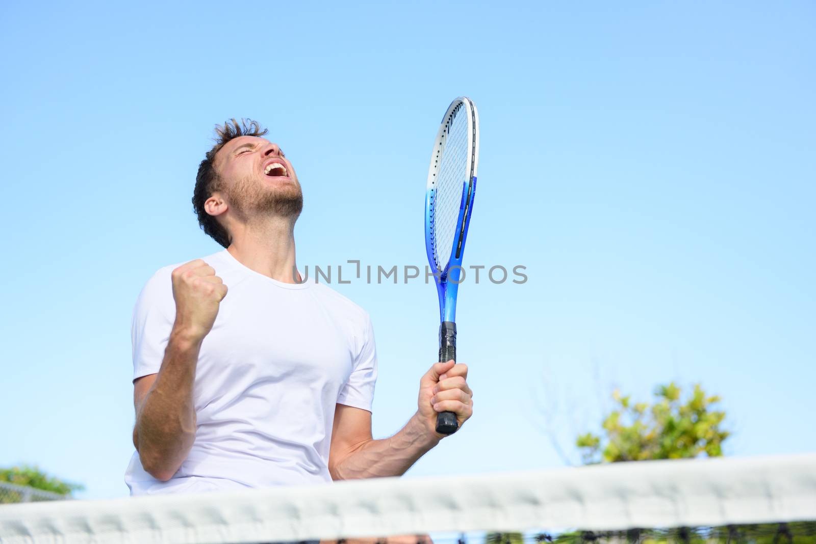 Tennis player man winning cheering celebrating victory. Winner man happy in celebration of success and win. Fit male athlete on tennis court outdoors holding tennis racket in triumph by the net.