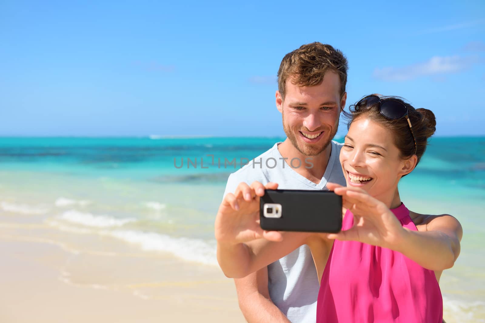 Smartphone - beach vacation couple taking selfie photograph using smartphone relaxing and having fun holding smart phone camera. Young beautiful multicultural Asian Caucasian couple on summer beach.