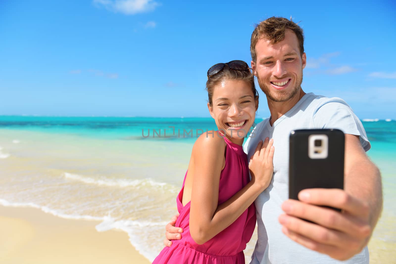 Beach vacation couple taking selfie photograph using smartphone relaxing and having fun holding smart phone camera. Young beautiful multicultural Asian Caucasian couple on summer beach.
