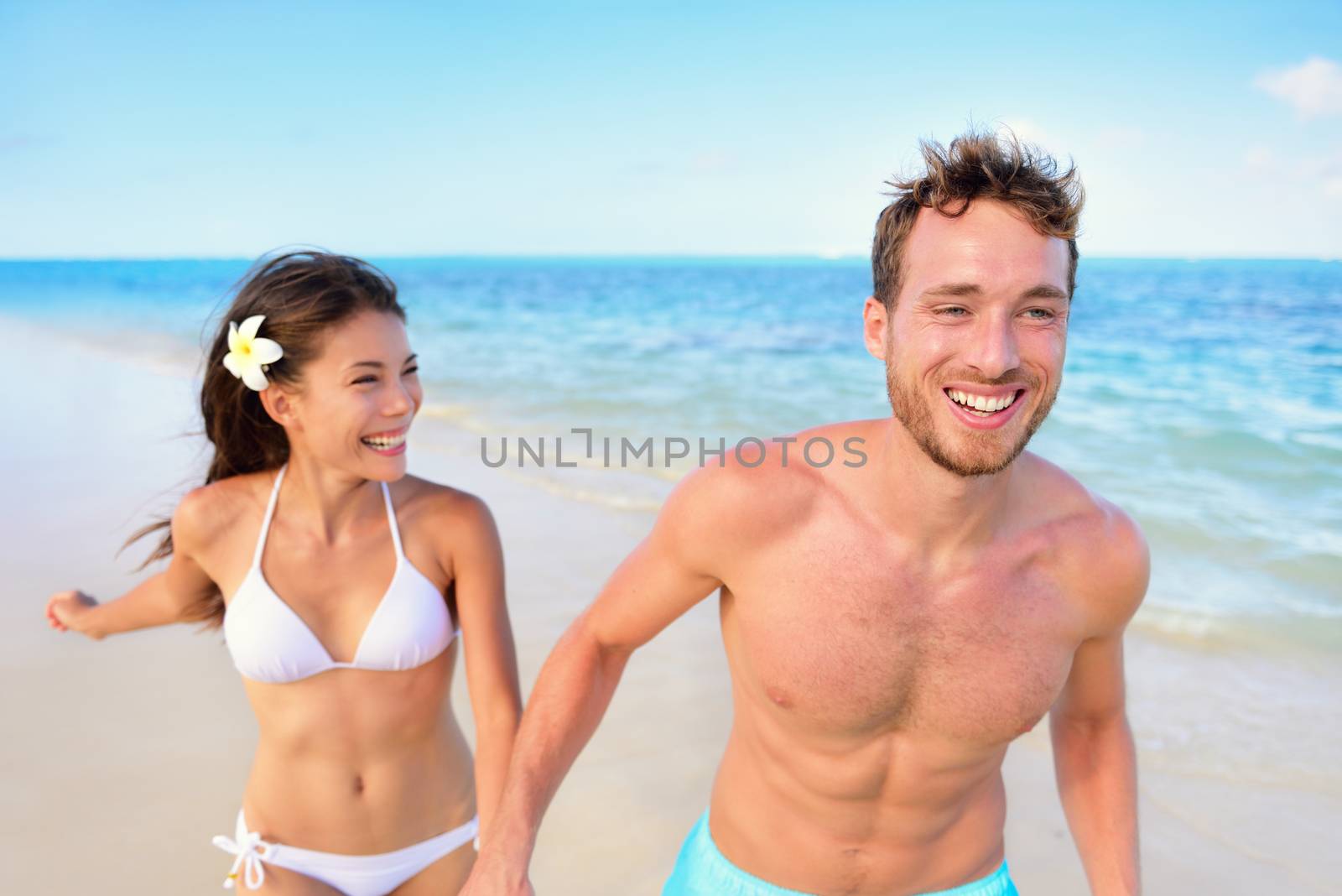 Beach fun - couple happy in love laughing running together on beach during vacation holidays travel. Mixed race asian chinese woman and Caucasian man holding hands in the sun.