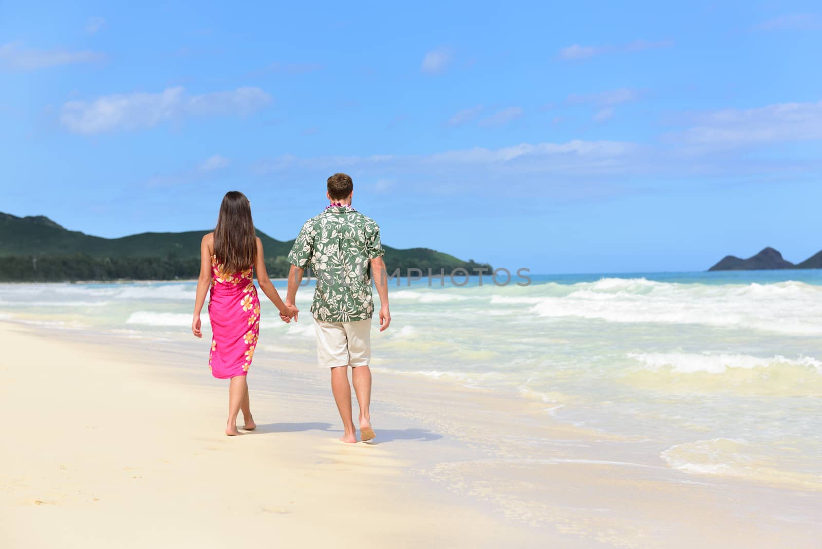 Hawaii honeymoon couple of newlyweds walking on tropical beach in Hawaiian apparel, pink sarong dress and green Aloha shirt for Polynesian cultural tradition. Young people holding hands happy in love.