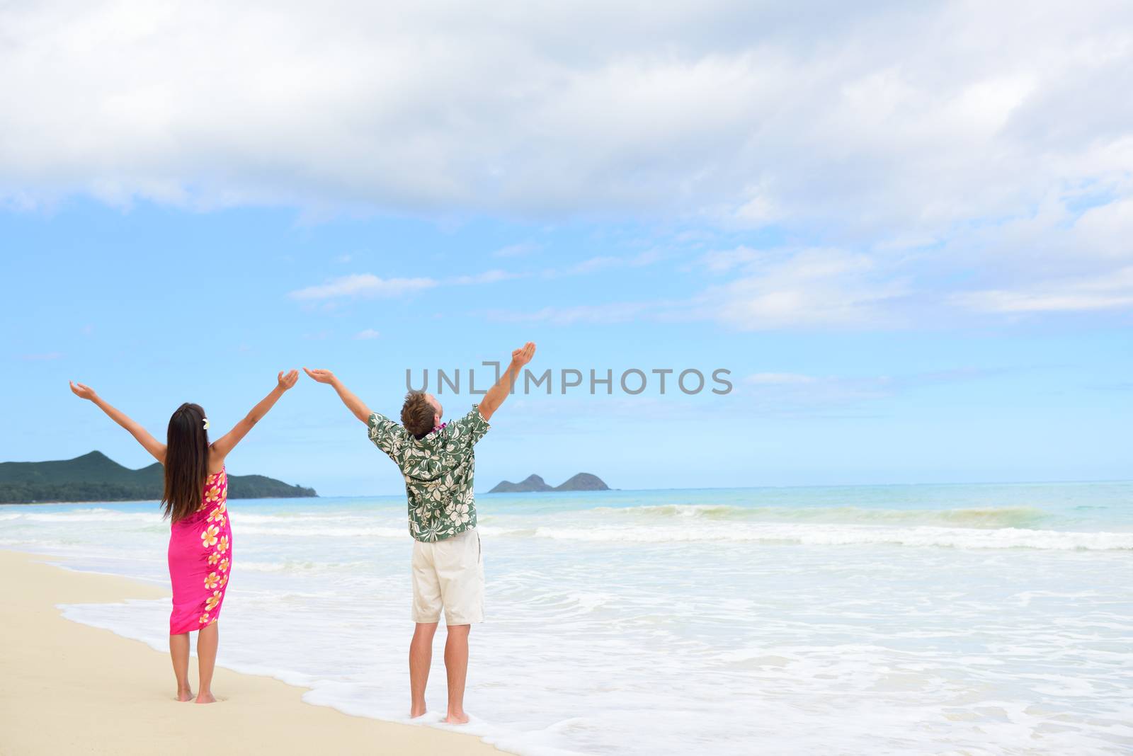 Happy freedom couple on Hawaiian beach vacations. Full length people standing with arms outstretched up to the sky showing happiness and success in Hawaii wearing traditional Aloha shirt and dress.