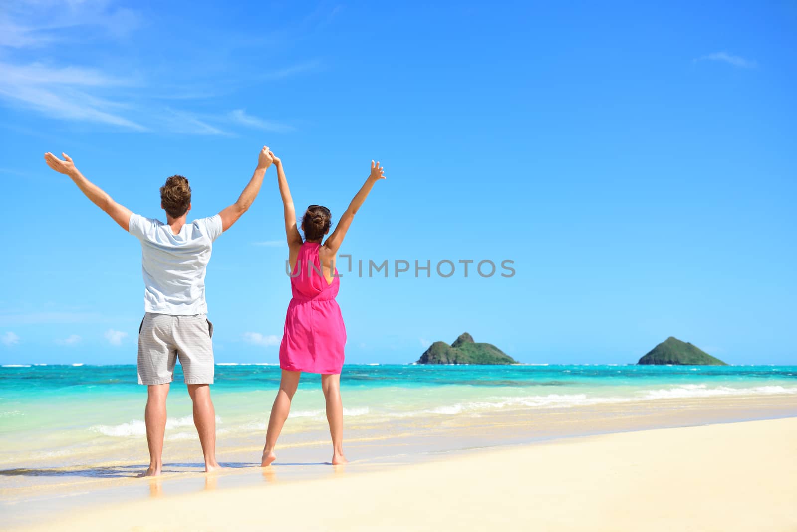 Summer beach happy free couple cheering on travel holiday. Back view of young people with arms raised to the sky showing success and freedom on Lanikai beach, Oahu, Hawaii, USA with Mokulua Islands.