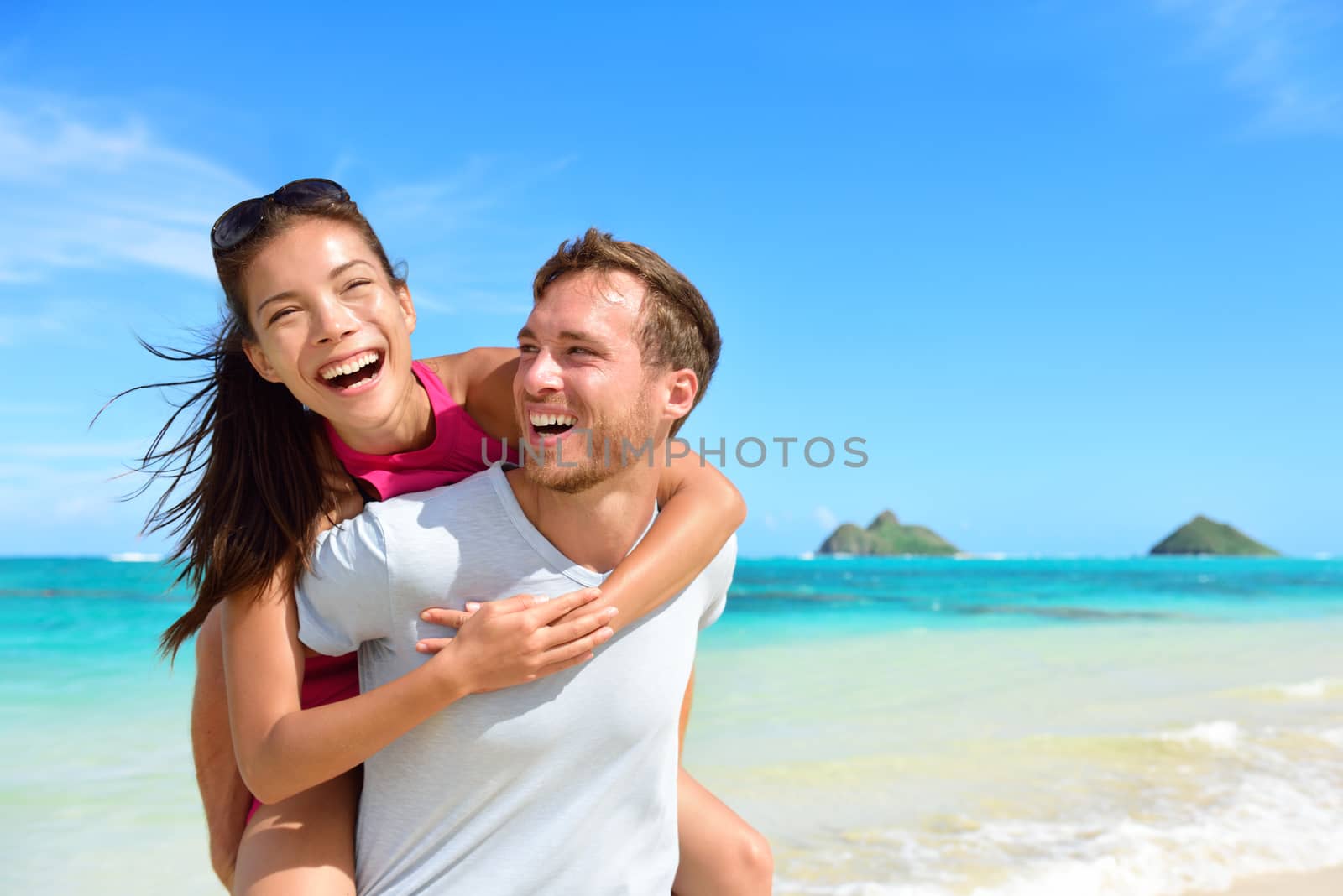 Beach couple having fun laughing on Hawaii holiday. Beautiful Asian mixed race woman piggybacking on Caucasian boyfriend on Lanikai beach, Oahu, Hawaii, USA with Mokulua Islands.