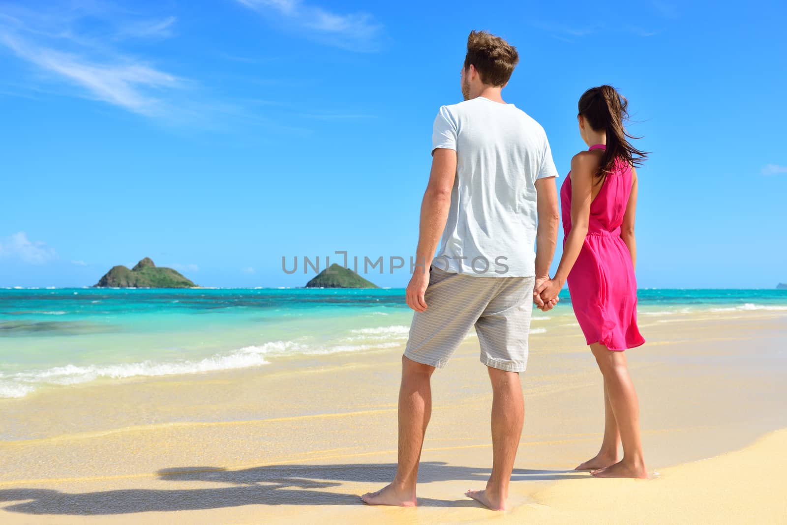 Beach couple looking at ocean view from behind. Couple standing on white sand in pink dress and beachwear on vacations on Lanikai beach, Oahu, Hawaii, USA with Na Mokulua Islands.