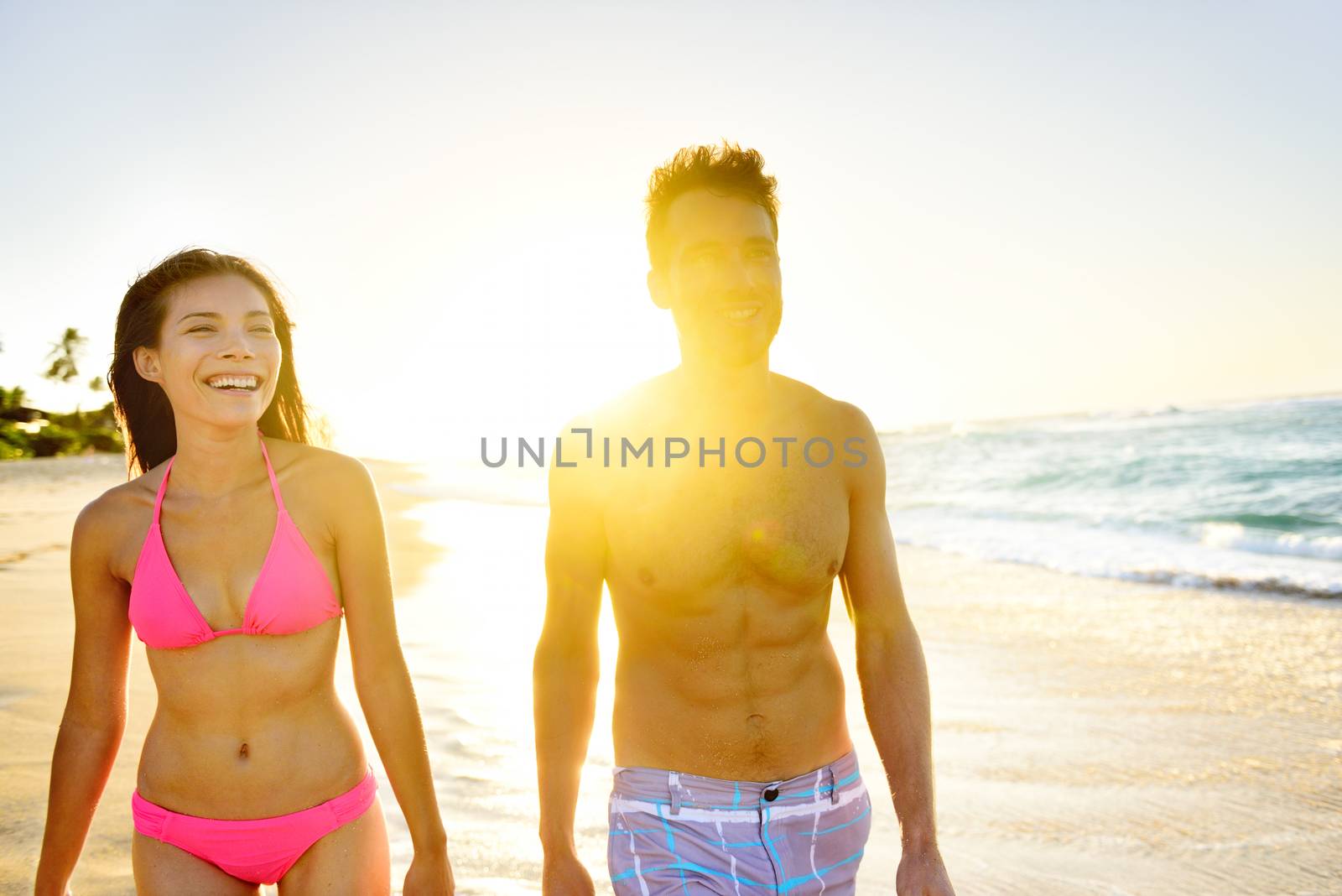Beach Lifestyle Couple Walking on Beach at Sunset enjoying healthy active lifestyle in summer sun. People in beachwear, woman in bikini and man in casual swim shorts.