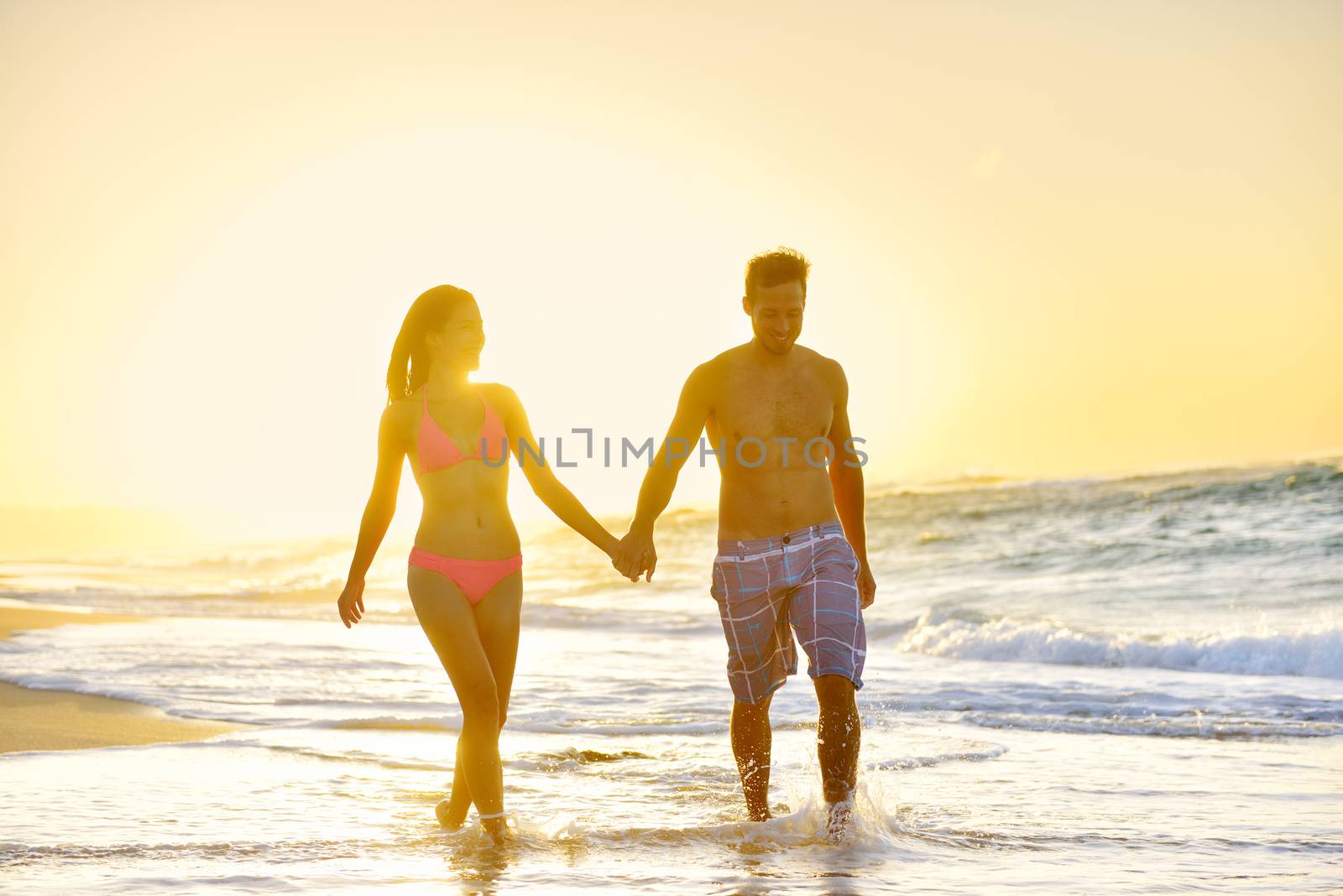 Romantic honeymoon couple in love holding hands walking on beautiful sunset at beach in waterfront. Lovers or newlywed married young couple by the sea enjoying vacation travel holiday. Oahu Hawaii USA