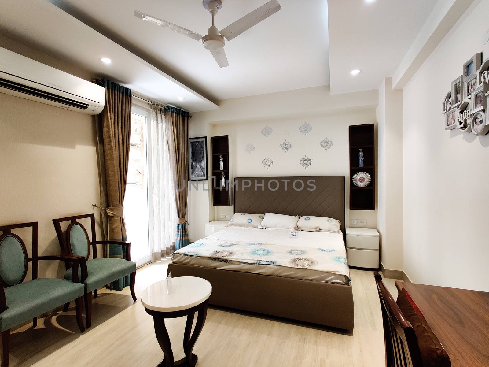 Wide angle shot of a modern large master bedroom with elegant bed, small coffee table and chairs and fan by Shalinimathur