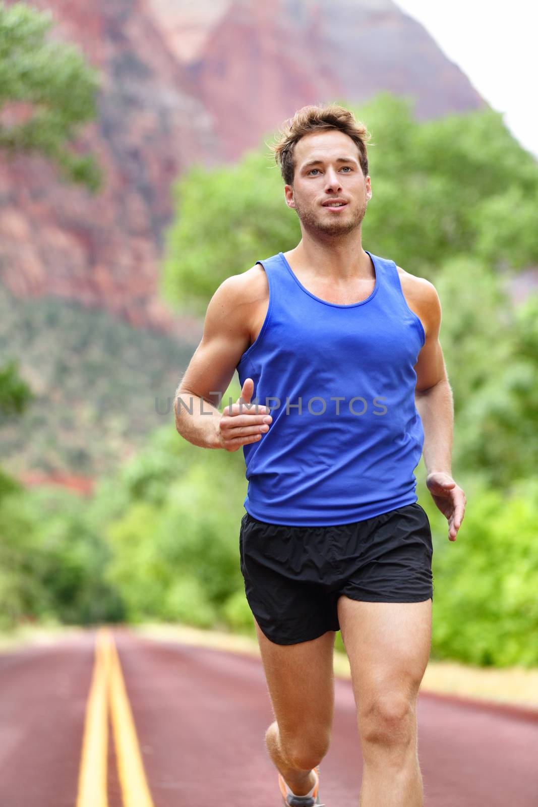 Sport and fitness runner man running on road training for marathon run doing sprint interval workout outdoors in summer. Male athlete sports model fit and healthy smiling with aspirations.