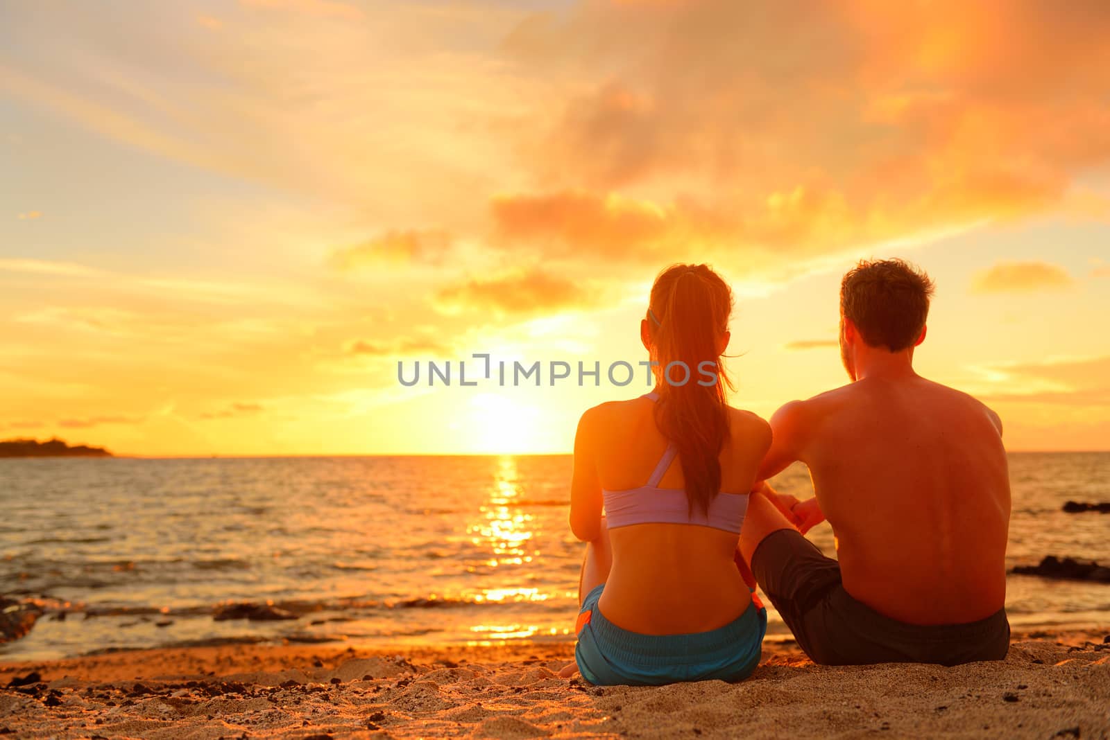 Happy Romantic Couple Enjoying Beautiful Sunset at the Beach Sitting in Sand looking at ocean sea and colorful yellow sky.