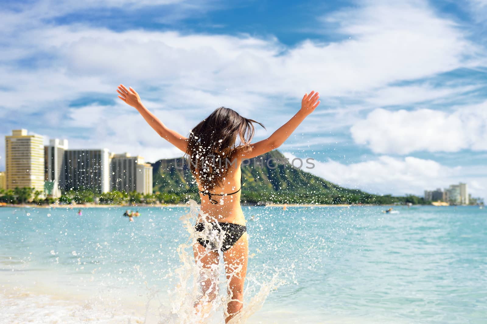 Beach fun - happy woman on Hawaii Waikiki vacation. Unrecognizable young adult from behind jumping of joy in water waves, arms up with diamond head mountain in the background, landmark of Honolulu.