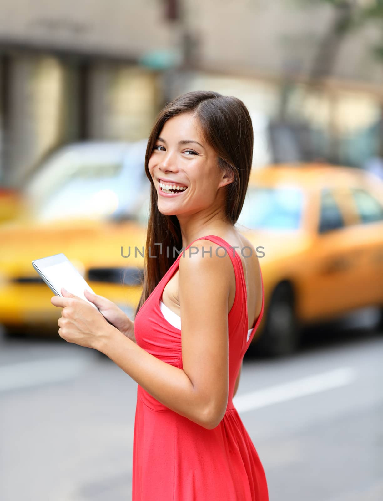 Woman in New York City using Tablet computer, standing in street with yellow taxi cab. Beautiful young woman in red dress smiling happy. Mixed race female model in Manhattan, New York City, USA.