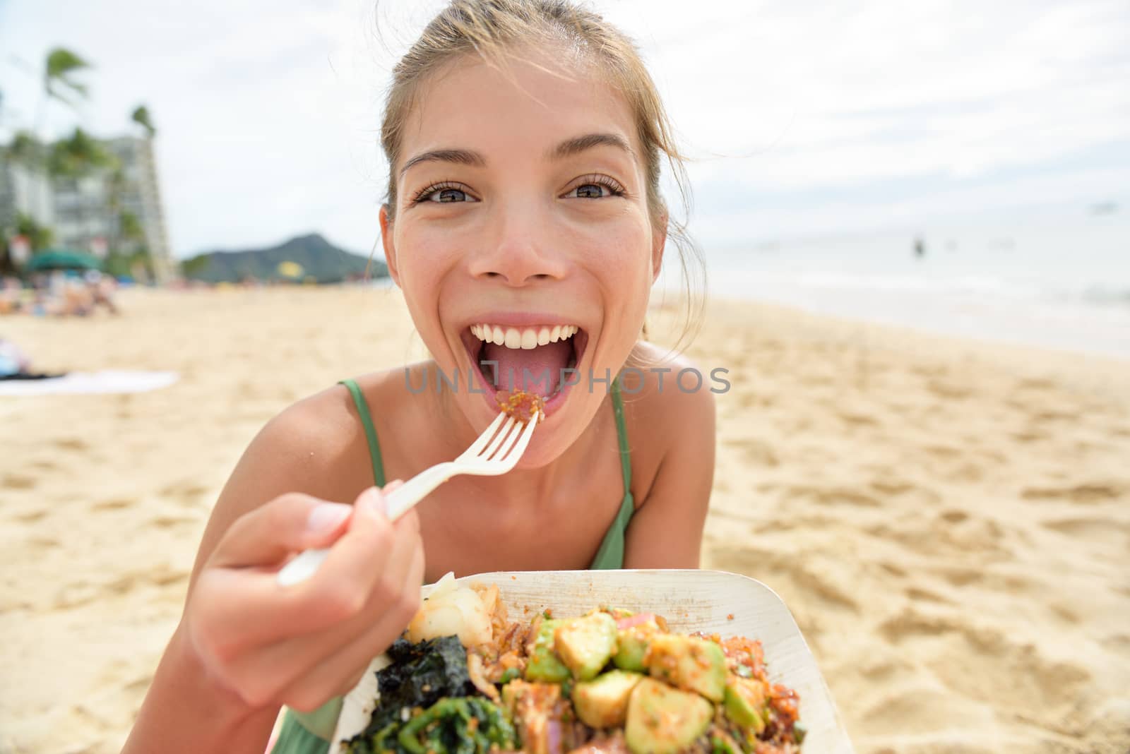 Funny woman eating salad healthy meal on beach in Hawaii. Asian Multiracial young female adult sitting at beach cafe eating a lunch during summer vacations in Waikiki, Honolulu, Hawaii, USA.