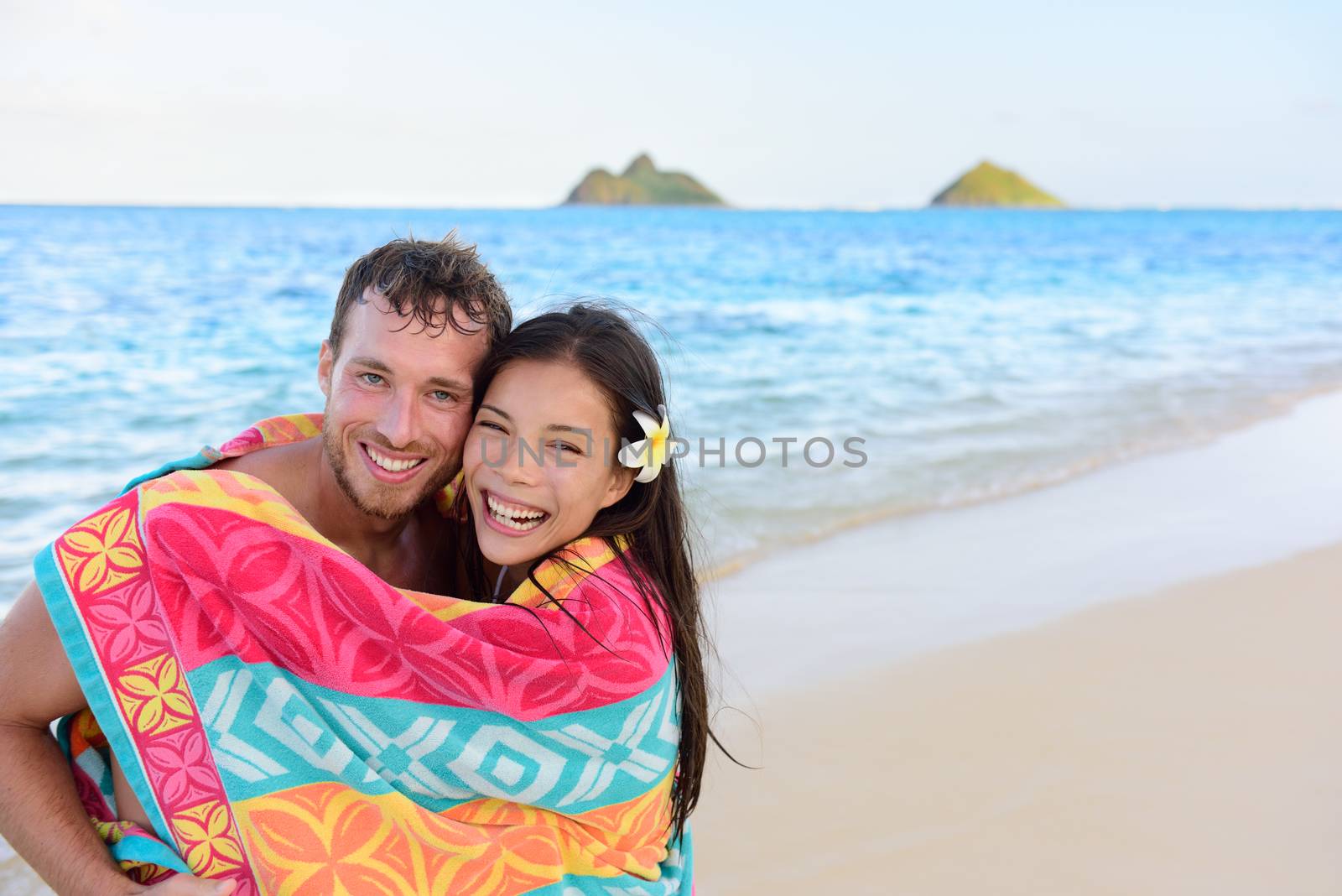 Swimming romantic couple wrapped in bathing towel on beach. Portrait of happy young interracial couple embracing each other having fun during holidays vacation travel. Asian woman, Caucasian man.