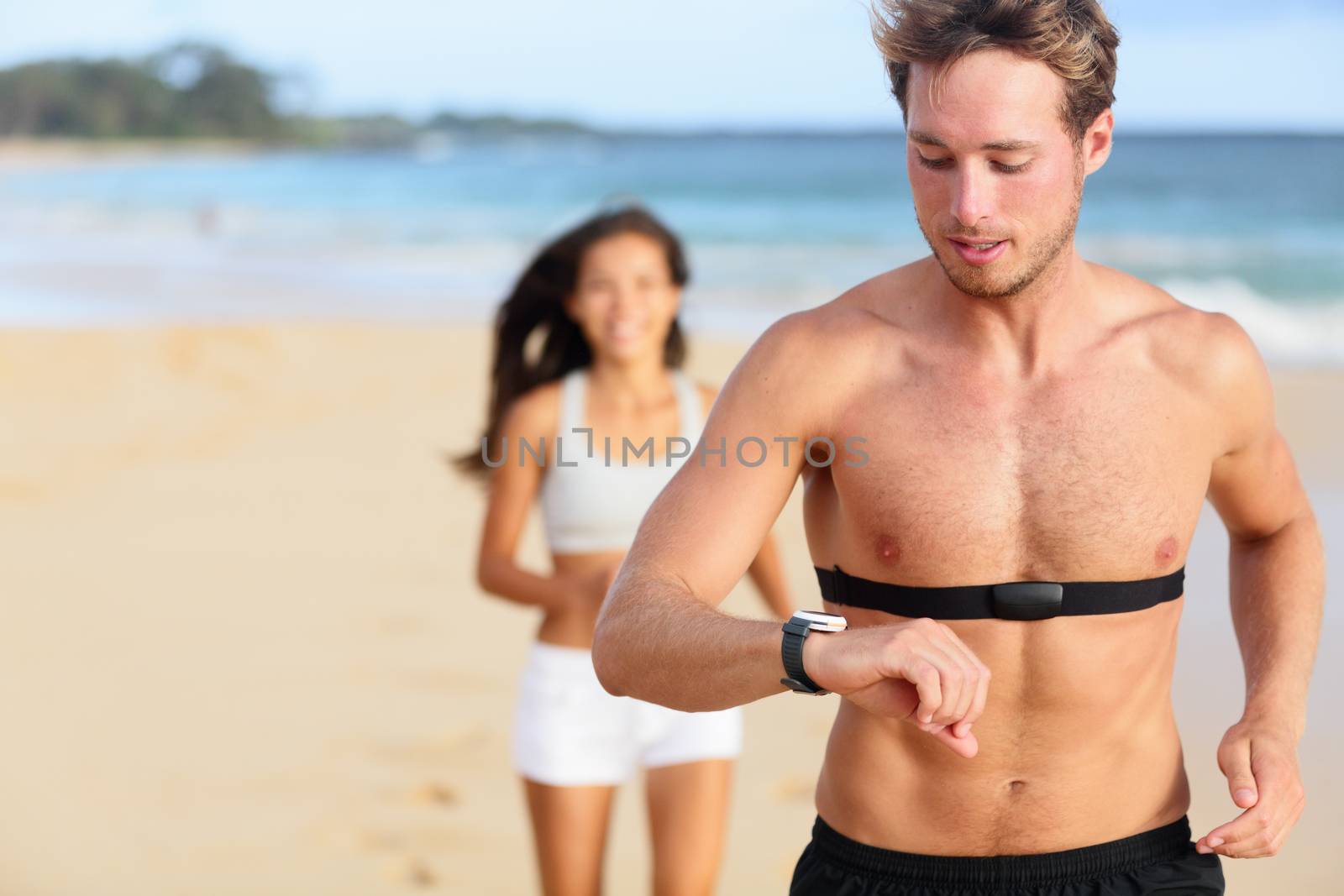 Running young man jogging on beach using heart rate monitor. Handsome shirtless male runner working out outside by the ocean wearing heart rate monitor. Closeup portrait of fit fitness athlete model.