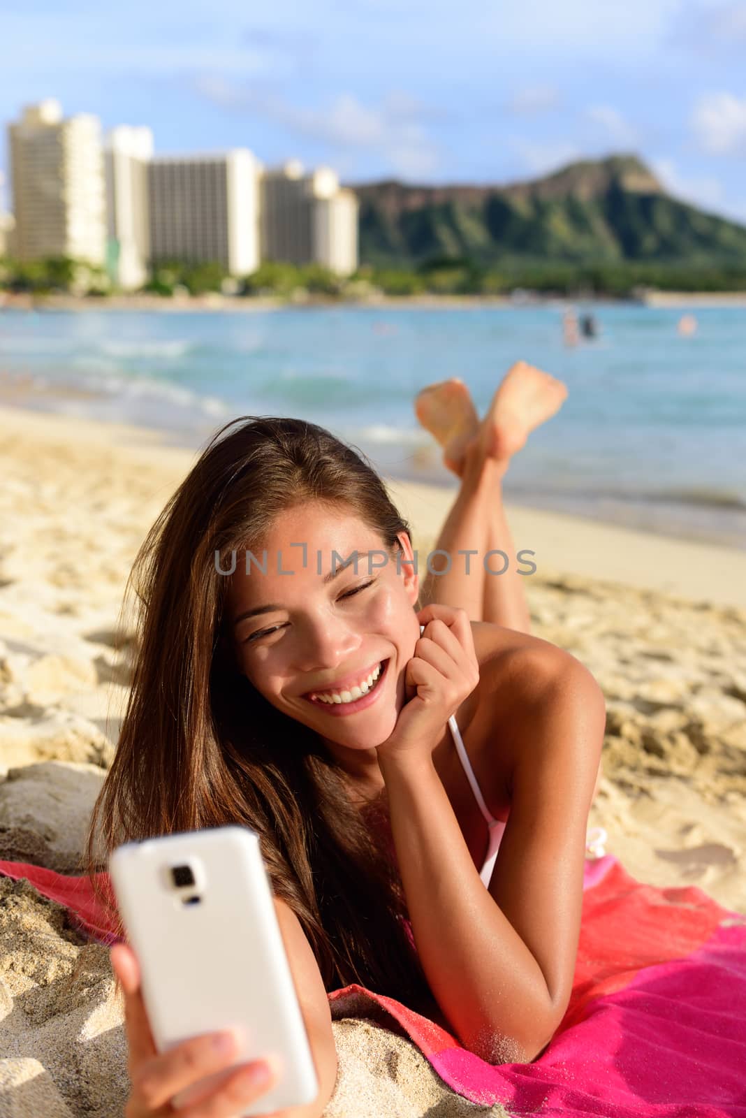 Smart phone woman using smartphone app on beach smiling laughing having fun. Girl reading or messaging or browsing on internet smiling happy outdoors. Mixed race female model on Waikiki Oahu, Hawaii.
