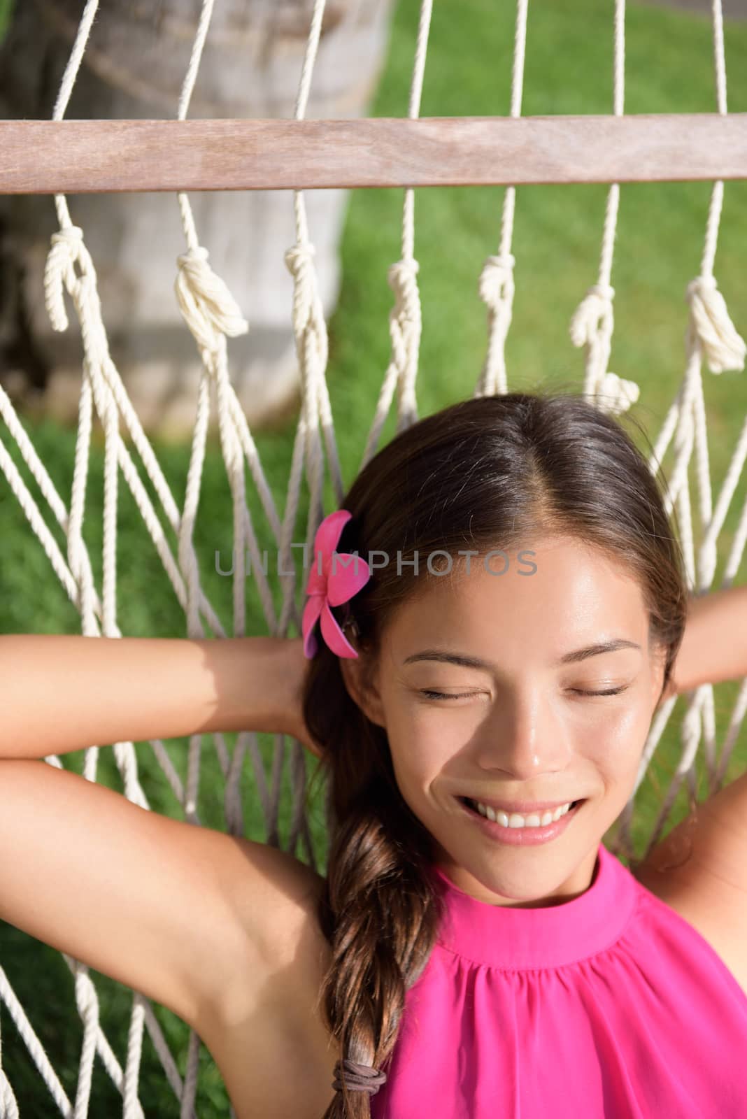 Happy relaxed young woman with hands behind head lying on hammock. Mixed race Asian / Caucasian female is smiling while dreaming. She is enjoying sunlight in park.