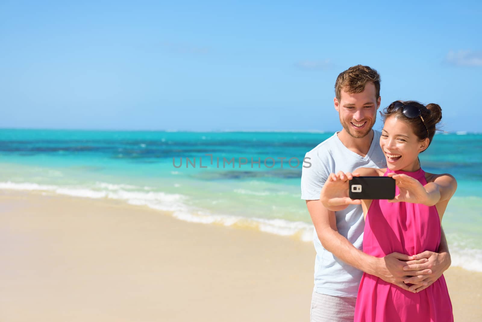 Happy young multiethnic couple taking selfie on mobile phone at beach. Loving partners are standing together while clicking pictures. Tourists are enjoying together at beach.
