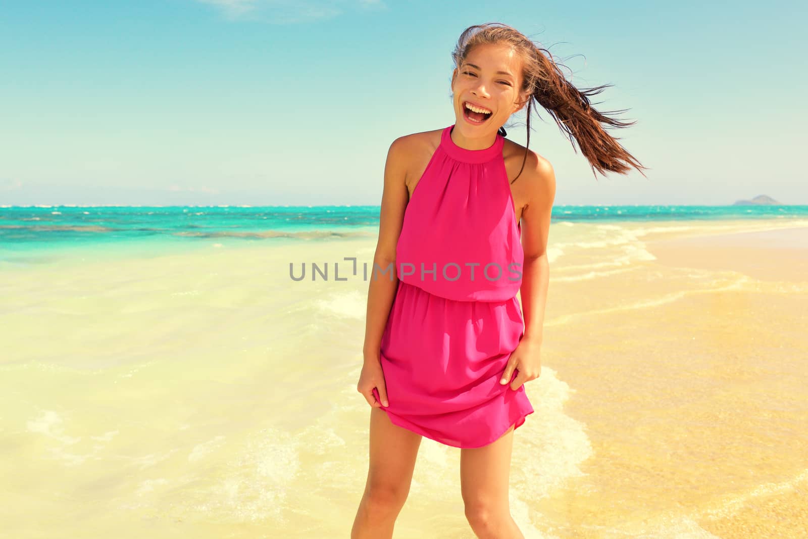 Portrait of happy young woman in pink dress standing at beach. Beautiful mixed race Asian / Caucasian female is enjoying her summer vacation. Carefree woman is laughing while standing on shore.