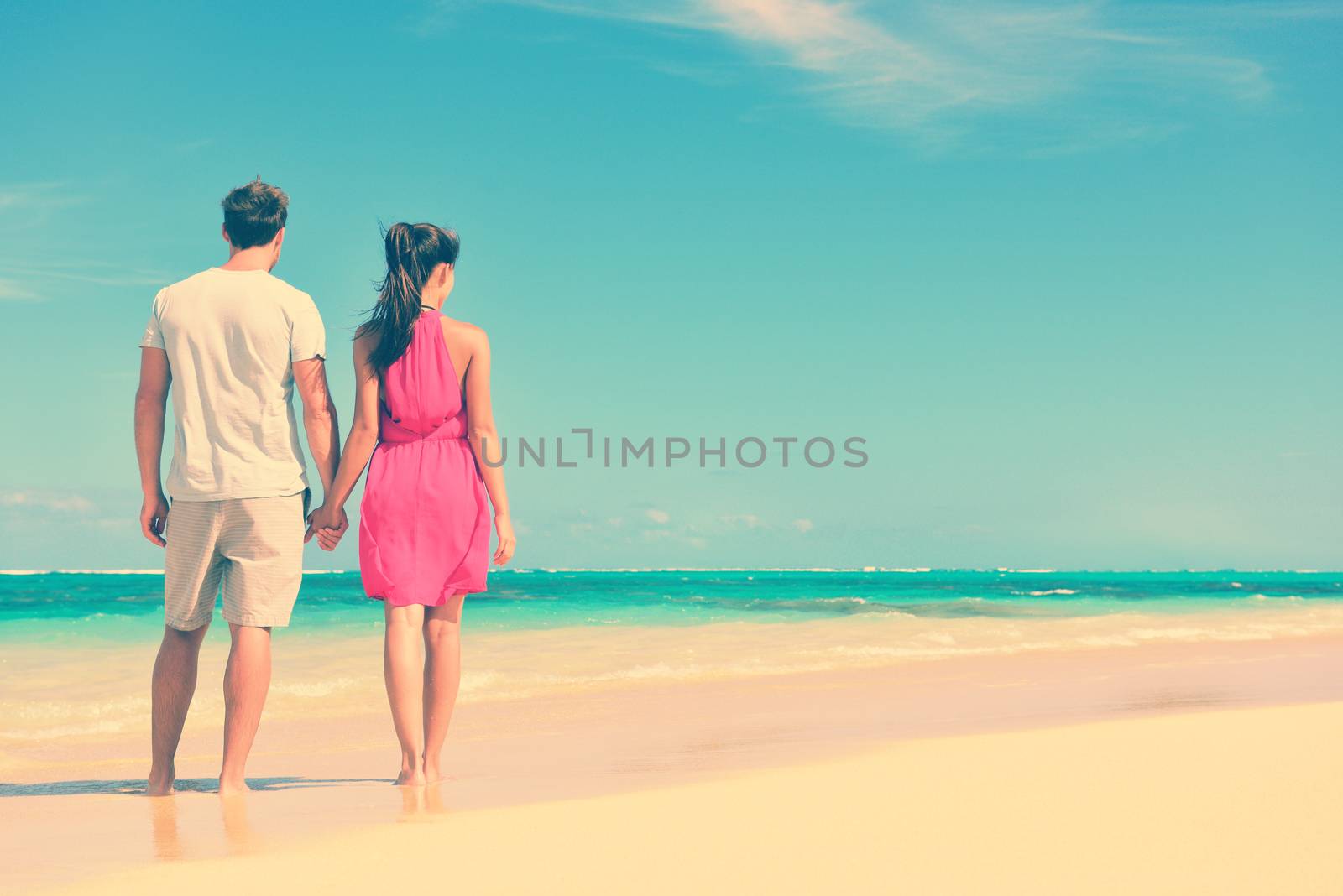 Couple standing on beach travel holding hands. Rear view of multiethnic romantic couple on shore. Young partners are watching sea. Tourists are enjoying their summer vacation at beach.