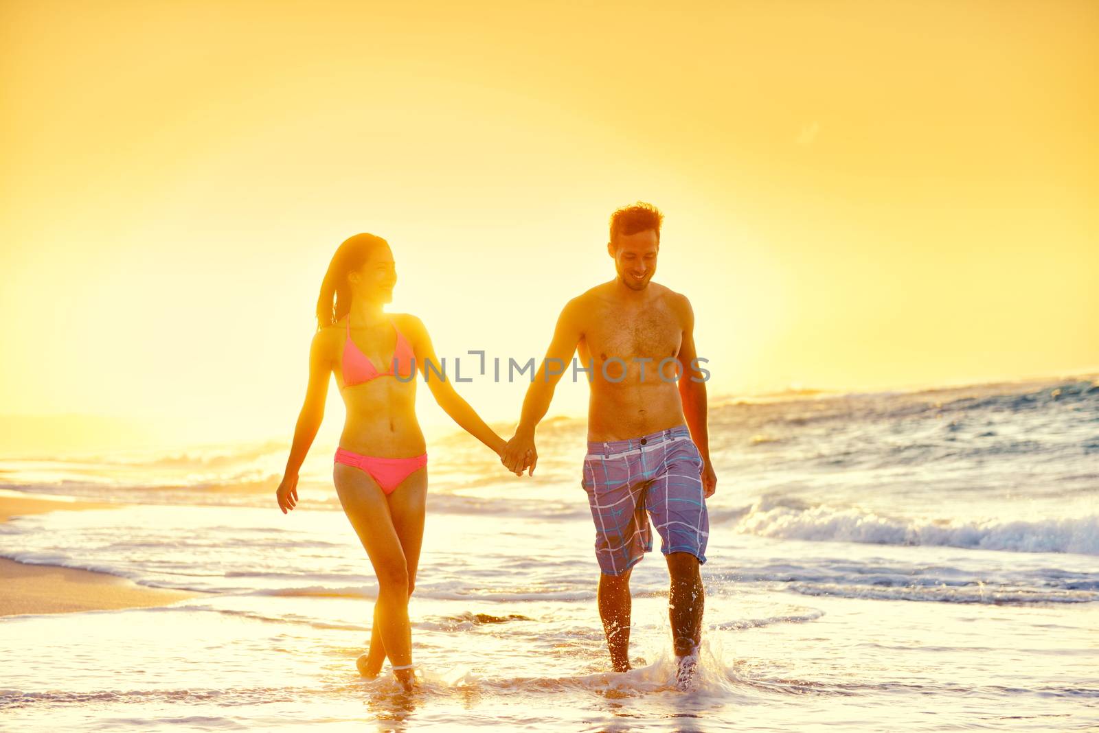 Young couple in swimwear walking at beach. Multiethnic romantic woman and man partners are holding hands while enjoying summer vacation. Loving tourists are enjoying sunset at beach.