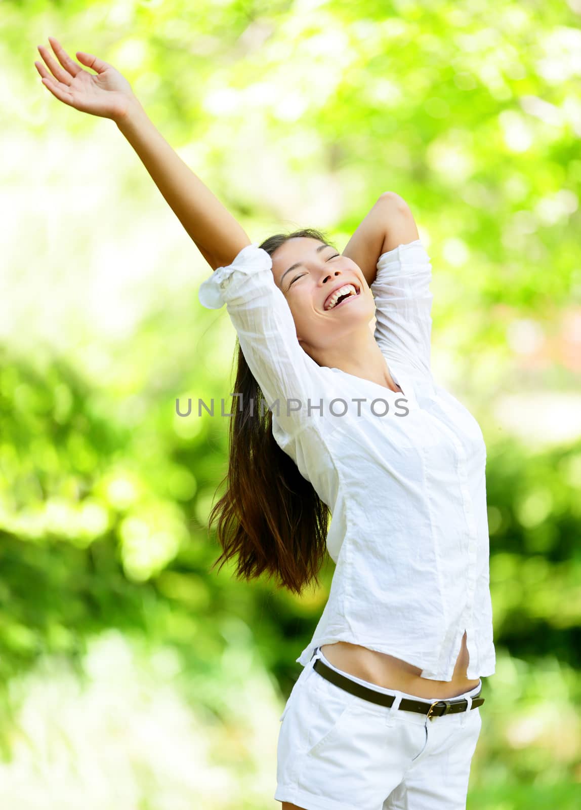 Happy young woman raising arms in park. Attractive mixed race Asian / Caucasian female is in casuals. She is closing eyes while smiling.