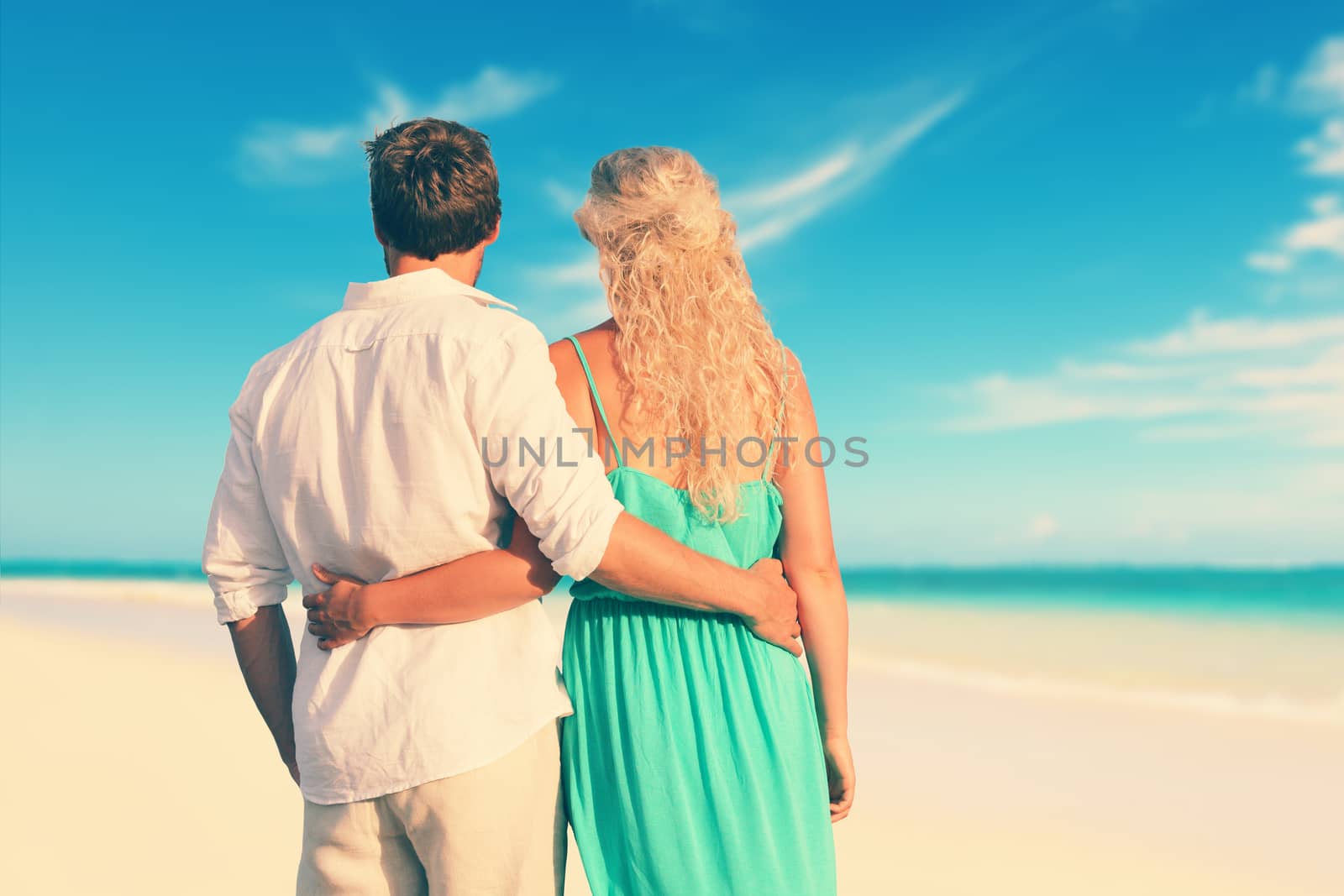 Rear view of young Caucasian couple with arms around. Loving partners are standing at shore. Man and woman are enjoying summer vacation at beach.