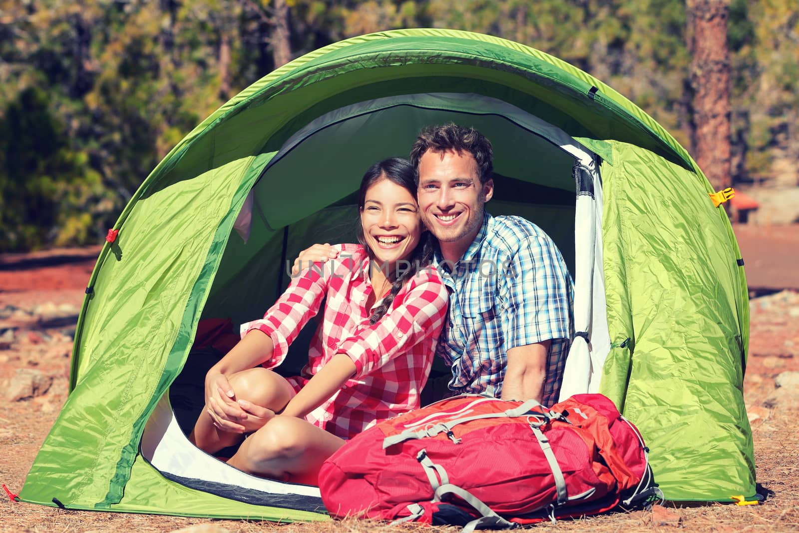 Happy young couple sitting in tent. Male and female hikers are resting in tent. Young partners are with hiking bag on sunny day.