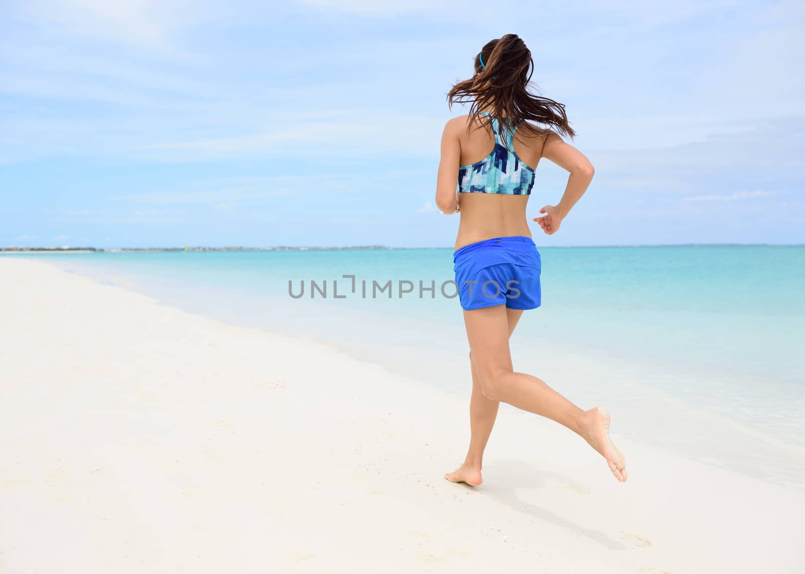 Runner training cardio running on beach. Back view of woman jogging barefoot in tropical destination barefoot in activewear blue sports bra and active shorts living a healthy life.