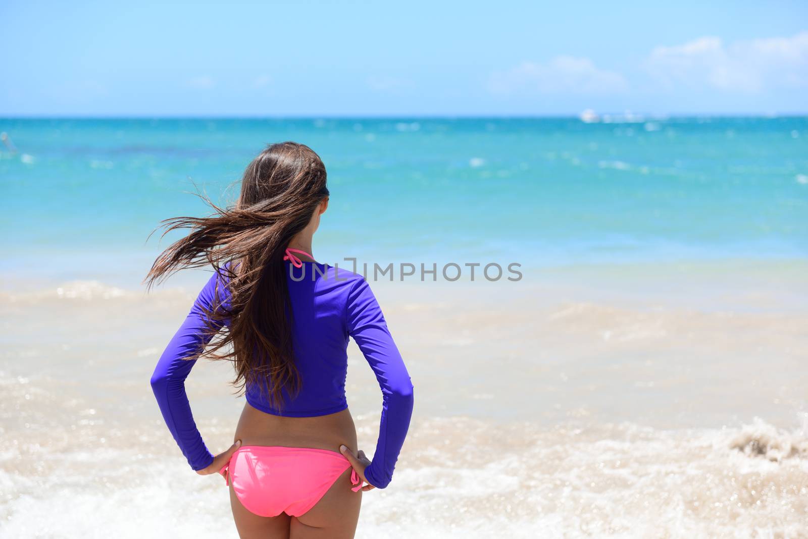 Beach girl going swimming in rashguard swimwear protective clothing. Woman standing in bikini and uv sun protection surf shirt looking at ocean waves. Healthy active lifestyle.