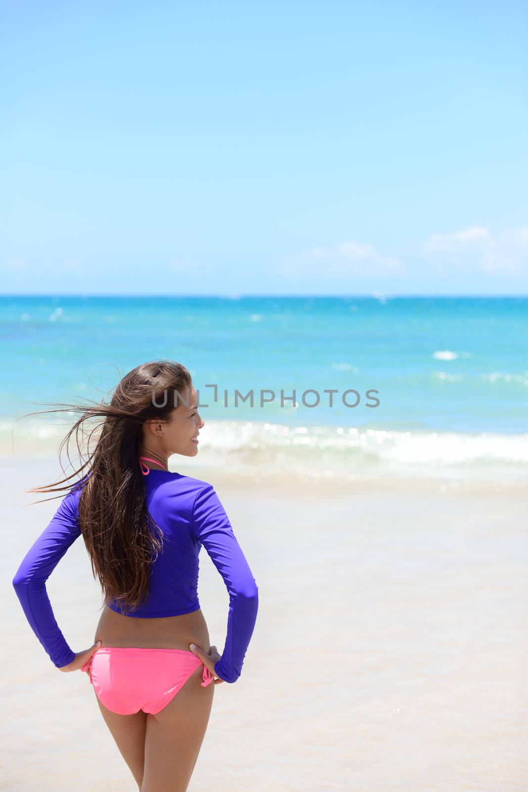 Woman relaxing on beach looking at the waves in sun protective swimwear t-shirt / rashguard to protect skin against rashes and uv rays during swimming or surfing.