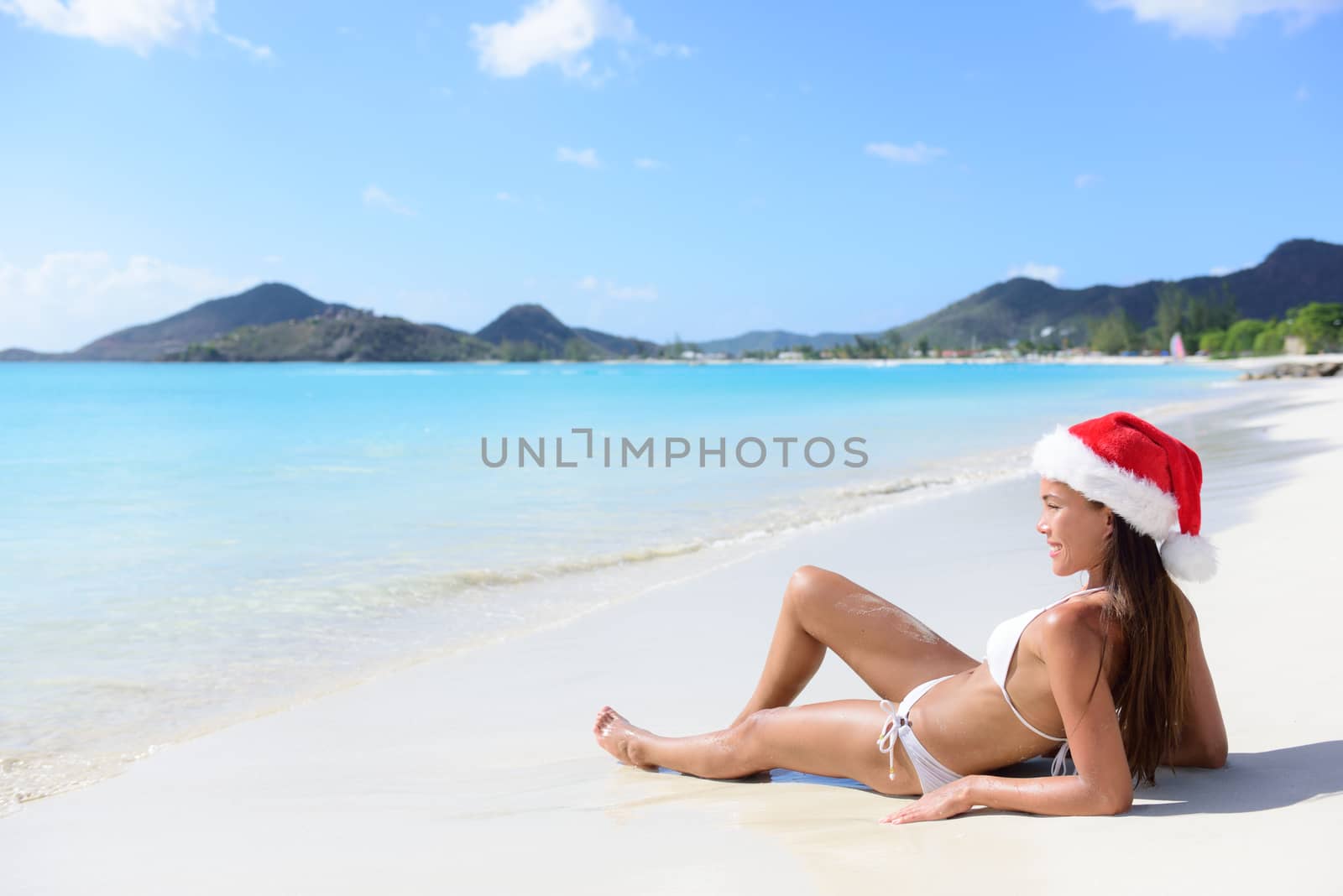 Christmas beach woman in santa hat and bikini on holidays travel vacation getaway relaxing on beautiful tropical beach with turquoise water in the Caribbean. Beautiful young female model sun tanning.
