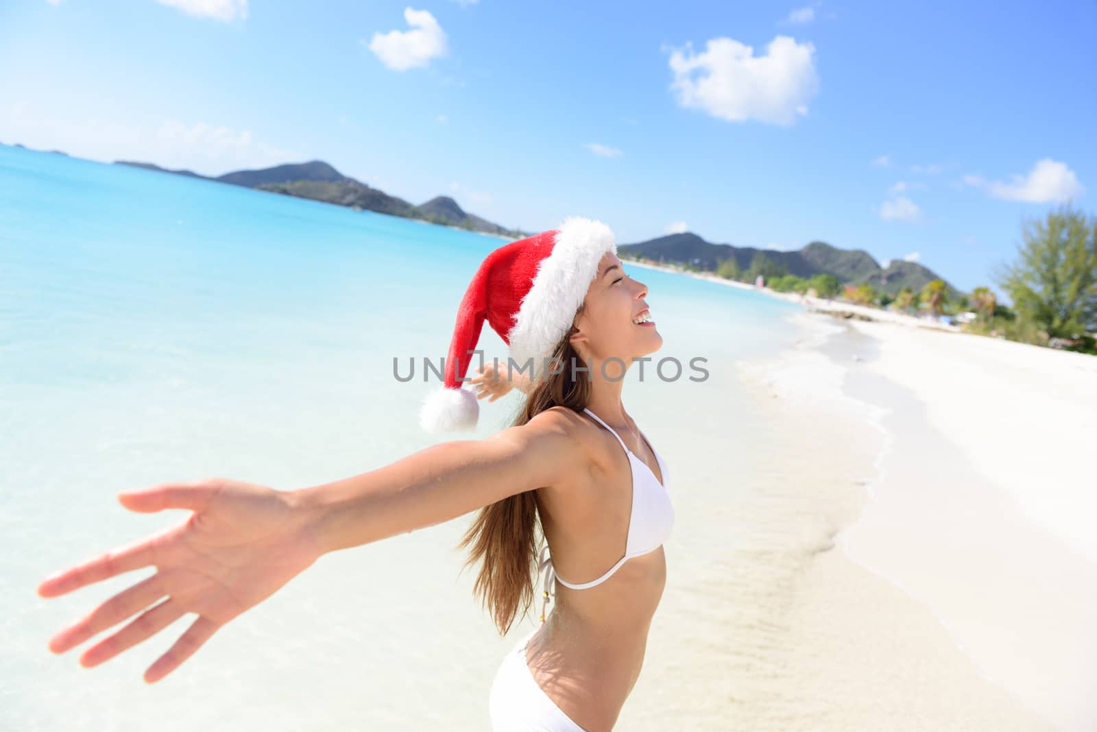 Christmas Santa hat bikini woman on beach vacation holiday getaway. Girl free and happy with arms outstretched of joy on tropical Caribbean beach. Beautiful girl in bikini having fun under the sun.