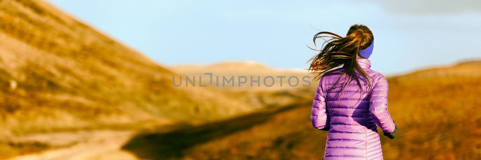 Running woman in autumn foliage background. Athlete runner on trail run outdoors panoramic banner. Active person jogging in down jacket from behind. by Maridav